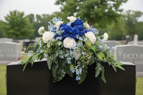 Handcrafted blue and white floral memorial saddle arrangement with navy carnations, white roses, light blue delicate blooms, and lush cascading ferns, displayed on a black granite headstone in a peaceful cemetery setting — custom made by GG's Handmade Creations in Scotch Village, Nova Scotia