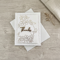 Gold foil daisy Thanks card with embossed white foliage on a softly styled wood and linen background.