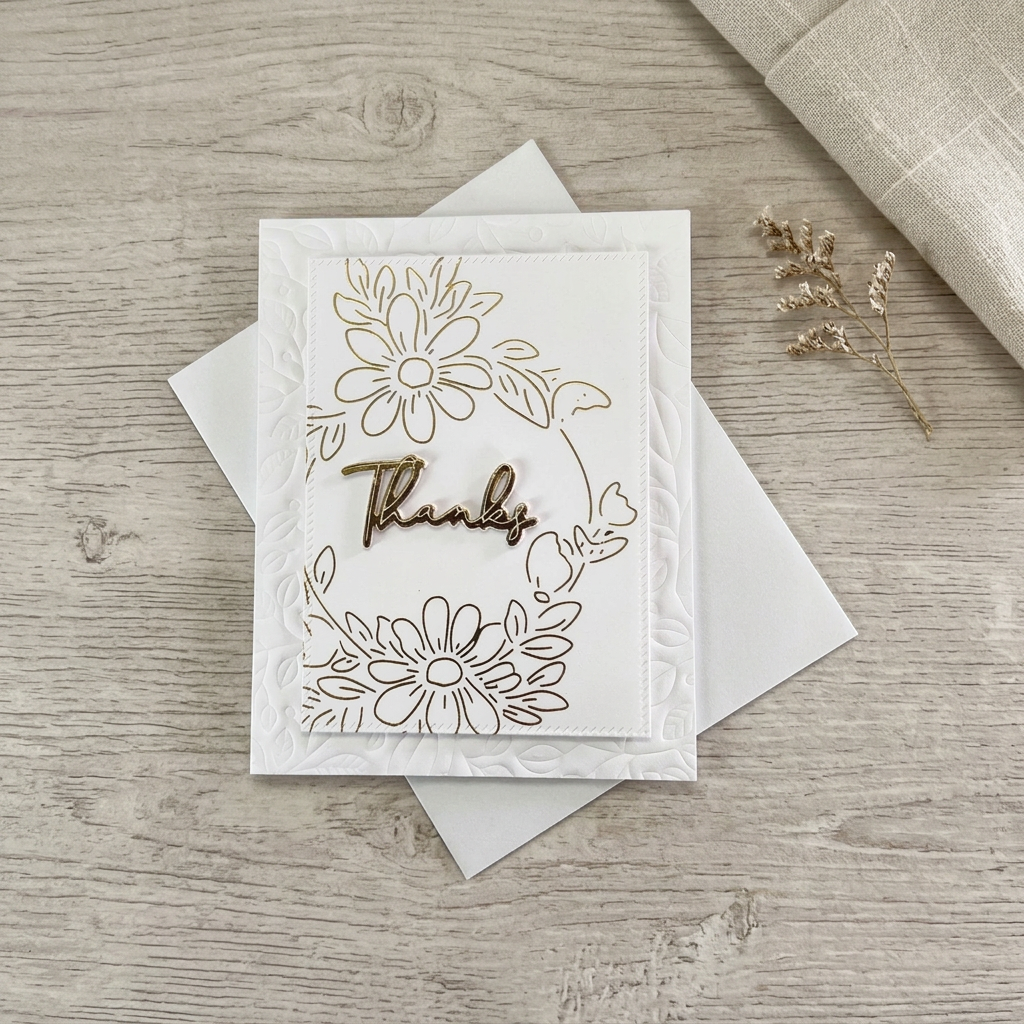 Gold foil daisy Thanks card with embossed white foliage on a softly styled wood and linen background.